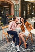 dois jovem à moda mulheres sentado às cafeteria, conversando, bisbilhotice, à moda na moda equipamento foto