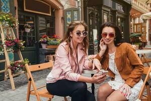 dois jovem à moda mulheres sentado às cafeteria, conversando, bisbilhotice, à moda na moda equipamento foto