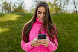 jovem sorridente mulher dentro Rosa suéter caminhando dentro verde parque usando telefone foto