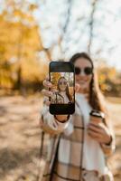 atraente jovem mulher caminhando dentro outono vestindo Jaqueta usando telefone foto