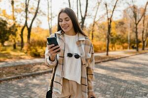 atraente jovem mulher caminhando dentro outono vestindo Jaqueta usando telefone foto