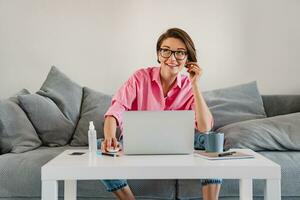 mulher dentro Rosa camisa sentado relaxado em sofá às casa às mesa trabalhando conectados em computador portátil foto