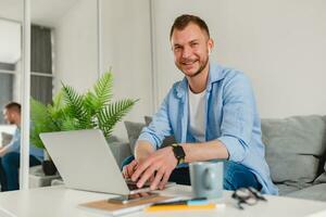 bonito sorridente homem dentro camisa sentado relaxado em sofá às casa às mesa trabalhando conectados em computador portátil foto