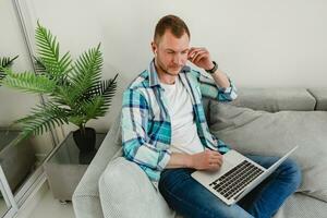 bonito sorridente homem dentro camisa sentado relaxado em sofá às casa às mesa trabalhando conectados em computador portátil foto