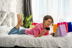 bonita feliz mulher deitado em cama às casa compras conectados foto