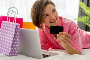 bonita feliz mulher deitado em cama às casa compras conectados foto