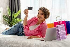 bonita feliz mulher deitado em cama às casa compras conectados foto