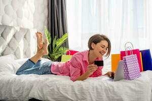 bonita feliz mulher deitado em cama às casa compras conectados foto