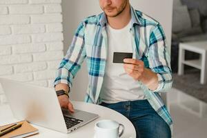 bonito sorridente homem dentro camisa sentado dentro cozinha às casa às mesa trabalhando conectados em computador portátil foto