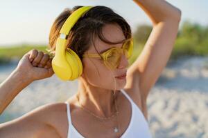 feliz sorridente mulher ouvindo para música dentro colorida amarelo fones de ouvido em ensolarado de praia dentro verão foto