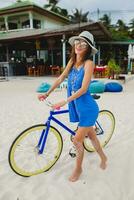 jovem atraente sorridente mulher dentro azul vestir caminhando em tropical de praia com bicicleta foto