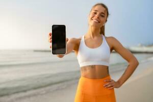 bonita jovem sorridente mulher fazendo Esportes dentro a manhã dentro à moda esporte equipamento roupa de esporte, segurando Smartphone tela fechar-se foto