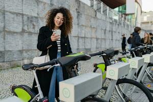 bonita encaracolado mulher alugando uma bicicleta dentro rua com a aplicativo foto