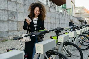 bonita encaracolado mulher alugando uma bicicleta dentro rua com a aplicativo foto