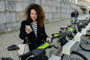 bonita encaracolado mulher alugando uma bicicleta dentro rua com a aplicativo foto