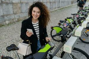 bonita encaracolado mulher alugando uma bicicleta dentro rua com a aplicativo foto