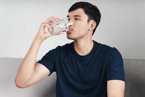 bonito homem asiático bebendo um copo d'água no sofá da sala foto