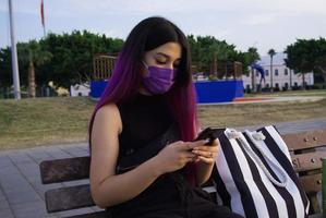 uma mulher de cabelo roxo com sacolas de compras. o celular na mão dela está descansando no parque. ela usa uma máscara roxa em seu rosto por causa do cobiçado 19.