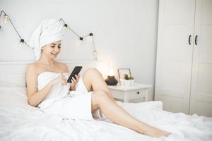 jovem mulher com uma toalha na cama branca mensagem de mensagens de texto no smartphone. foto