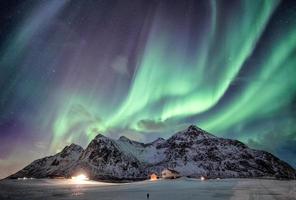 aurora boreal com cordilheira estrelada sobre neve com casa de iluminação em flakstad, ilhas lofoten, noruega