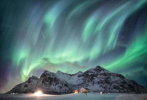 aurora boreal com cordilheira estrelada sobre neve com casa de iluminação em flakstad, ilhas lofoten, noruega foto