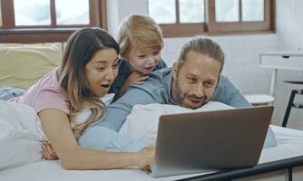 caucasiano família do três usando computador portátil enquanto deitado em cama junto, navegando Internet ou assistindo filme. foto