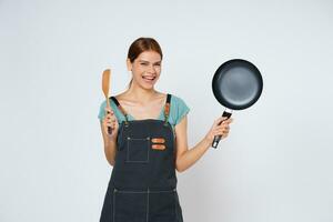 jovem mulher vestindo cozinha avental cozinhando e segurando panela e espátula isolado em branco fundo. foto