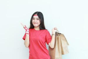 retrato do lindo ásia mulher vestindo vermelho equipamento a comemorar Indonésia independência dia de gesticulando carregando grande quantidade do compras bolsas com feliz expressão foto