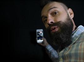 homem estiloso com barba e bigode olha para o telefone e faz uma selfie em um fundo preto