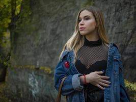 menina linda modelo loira com uma jaqueta jeans está de pé na rua contra um muro de pedra no parque foto