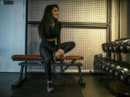 menina morena atraente com um top preto e legging sentada em um banco em uma sala de classificação em uma prateleira com halteres foto