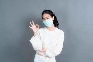 jovem mulher asiática usando máscara médica e mostrando sinal de ok