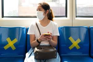 jovem mulher asiática usando máscara facial, usando um telefone inteligente e sentado no trem do céu. ela mantém o distanciamento social de outras pessoas durante o covid-19 ou surto de coronavírus foto