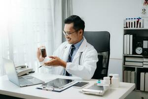 confiante jovem ásia masculino médico dentro branco médico uniforme sentar às escrivaninha trabalhando em computador. sorridente usar computador portátil escrever dentro médico Diário dentro clínica. foto