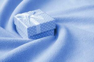 uma pequeno presente caixa com uma pequeno arco mentiras em uma cobertor do suave e peludo fantasma clássico azul cor lã tecido com uma muitos do alívio dobras. embalagem para uma presente para seu adorável namorada foto