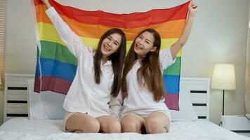 retratos do feliz ásia lésbica casal sentado com arco Iris bandeira sentado cama ao lado a janela. alegre dois jovem adulto mulheres vivo juntos com relação conceito. lgbtq estilo de vida orgulho mês foto