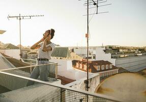 feliz jovem mulher em pé em telhado às pôr do sol ouvindo para música com fones de ouvido foto