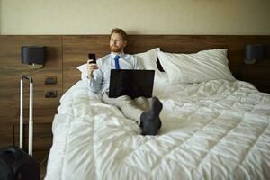 homem de negocios deitado em cama dentro hotel quarto usando computador portátil e célula telefone foto