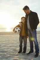 pai abraçando filho com futebol em a de praia às pôr do sol foto