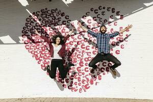 feliz jovem casal pulando dentro frente do uma tijolo parede com uma coração foto