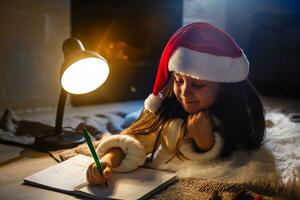 pequeno menina dentro santa chapéu escreve carta para santa claus perto Natal árvore e relógio foto