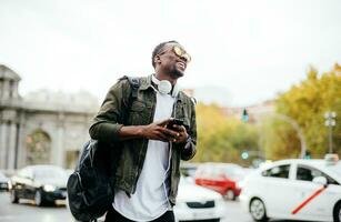 feliz homem vestindo oculos de sol segurando inteligente telefone enquanto em pé contra céu dentro cidade foto