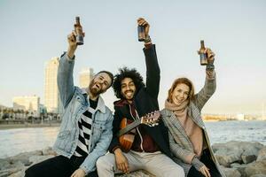 retrato do três feliz amigos com guitarra levantando Cerveja garrafas às a costa pôr do sol foto