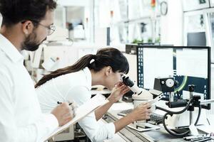 fêmea o negócio profissional olhando através microscópio enquanto em pé de masculino colega às laboratório foto