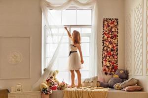 retrato de uma jovem com um vestido branco, endireitando as cortinas brancas claras perto da janela.