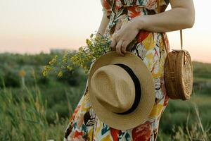 fechar-se detalhes do jovem mulher dentro verão vestir com rattan saco segurando Palha chapéu caminhando em pôr do sol dentro natureza. conceito do férias, viagem, verão estilo equipamento foto