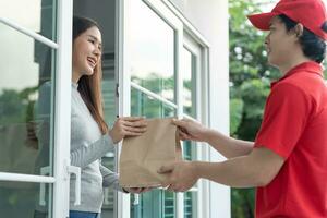 mulher asiática sorridente feliz recebe pacote de saco de papel de comida da casa da frente do correio. entregador enviar entrega expresso. compras online, recipiente de papel, levar, carteiro, serviço de entrega, pacotes foto