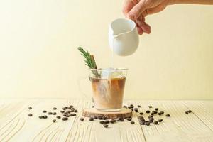 Derramando leite em copo de café preto com cubo de gelo, canela e alecrim no fundo de madeira foto