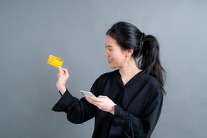 retrato de uma jovem asiática feliz mostrando um cartão de crédito de plástico enquanto segura o telefone celular em um fundo cinza foto