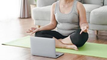 madura asiática gorda gordinha sentada no chão na sala de estar prática aula de ioga on-line com o computador. mulher tendo aula de treinamento de meditação no laptop. foto